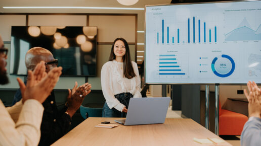 A meeting takes place in a modern office environment. A young woman has completed a successful presentation using a laptop and a huge computer monitor / television. Her colleagues giver her a round of applause for a job well done after creating a last-minute presentation. 